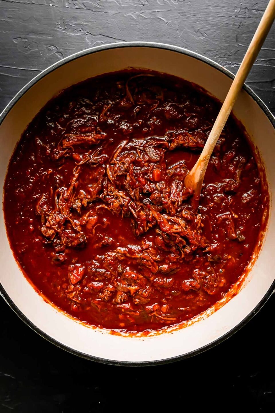 Beef ragu sauce fills a large white pot that sits atop a black textured surface. A wooden spoon rests inside of the pot for stirring the beef ragu recipe.
