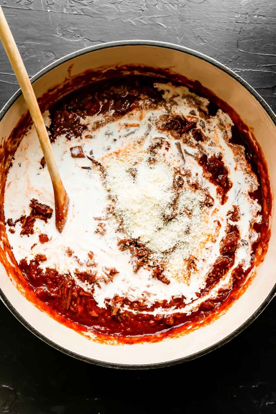 Beef ragu sauce fills a large white pot that sits atop a black textured surface. The sauce has just been finished with heavy cream and grated parmesan. A wooden spoon rests inside of the pot for stirring.