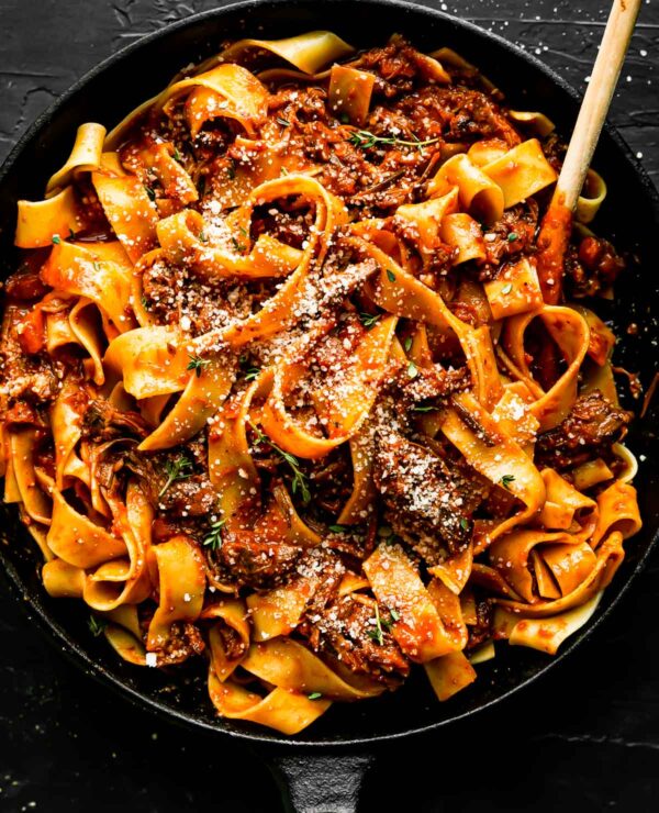 Finished beef ragu fills a large black skillet. The ragu pasta has been garnished with fresh thyme leaves and grated parmesan. The skillet sits atop a black textured surface and a wooden spoon rests inside of the skillet.