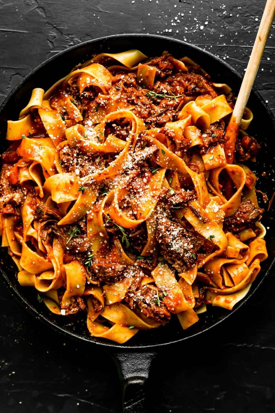 Finished beef ragu fills a large black skillet. The ragu pasta has been garnished with fresh thyme leaves and grated parmesan. The skillet sits atop a black textured surface and a wooden spoon rests inside of the skillet.