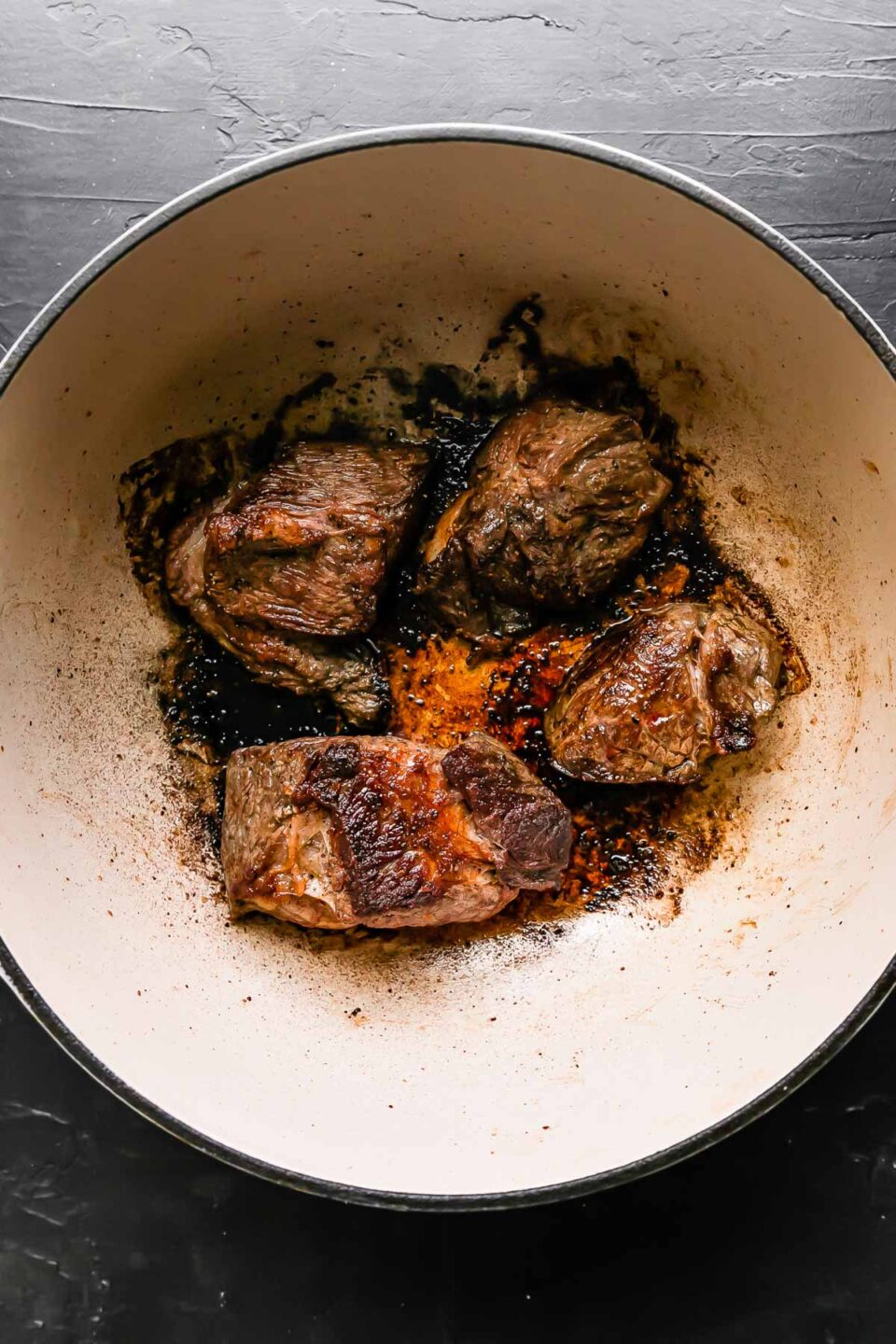 How to make braised beef ragu, step 1: Brown the beef. Browned beef chuck roast sections rest in the bottom of a large white pot that sits atop a black textured surface.