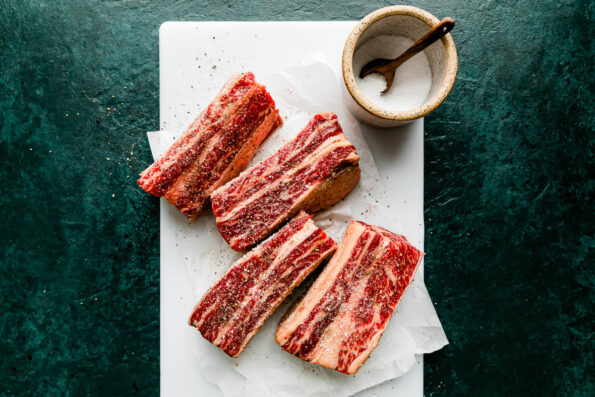 English-cut beef short ribs arranged atop a piece of parchment paper that sits atop a plastic cutting board atop a dark green textured surface. A small ceramic bowl filled with kosher salt rests atop the cutting board with a wooden spoon resting inside.