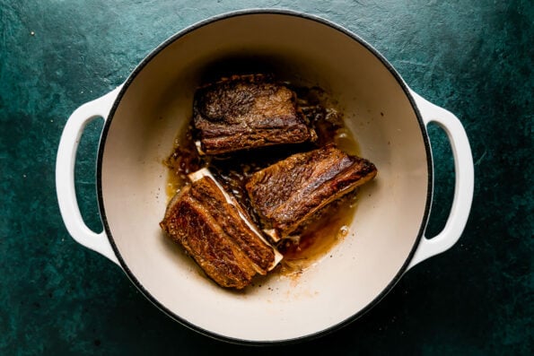 Browned English-cut beef short ribs rest inside of a white double-handled ceramic braising pot. The pot sits atop a dark green textured surface.