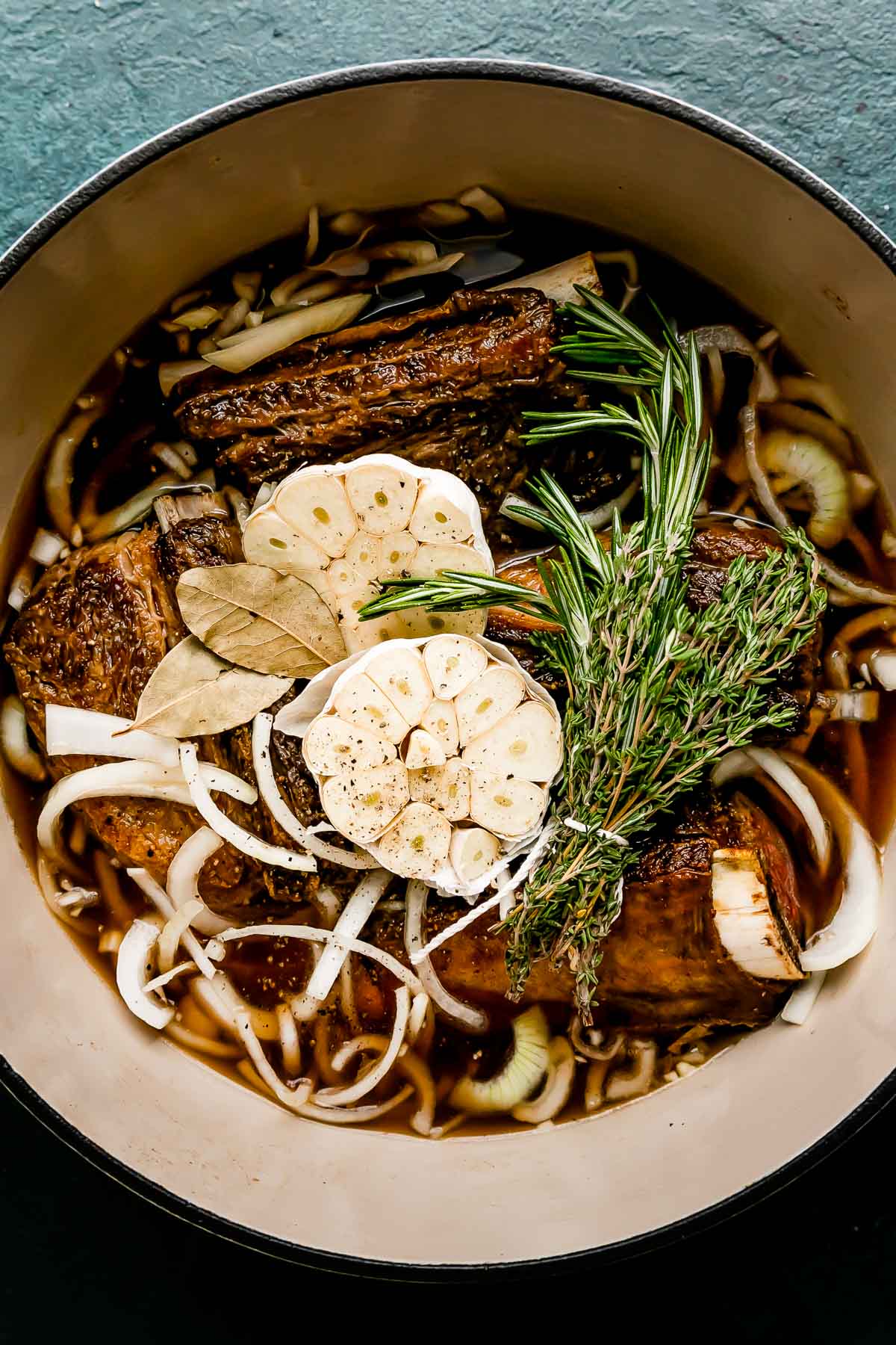 Browned short ribs rest inside of a white ceramic pot filled with braising liquid, garlic, sliced onions, bay leaves, and a bundle of fresh rosemary and thyme. The pot sits atop a dark green textured surface.
