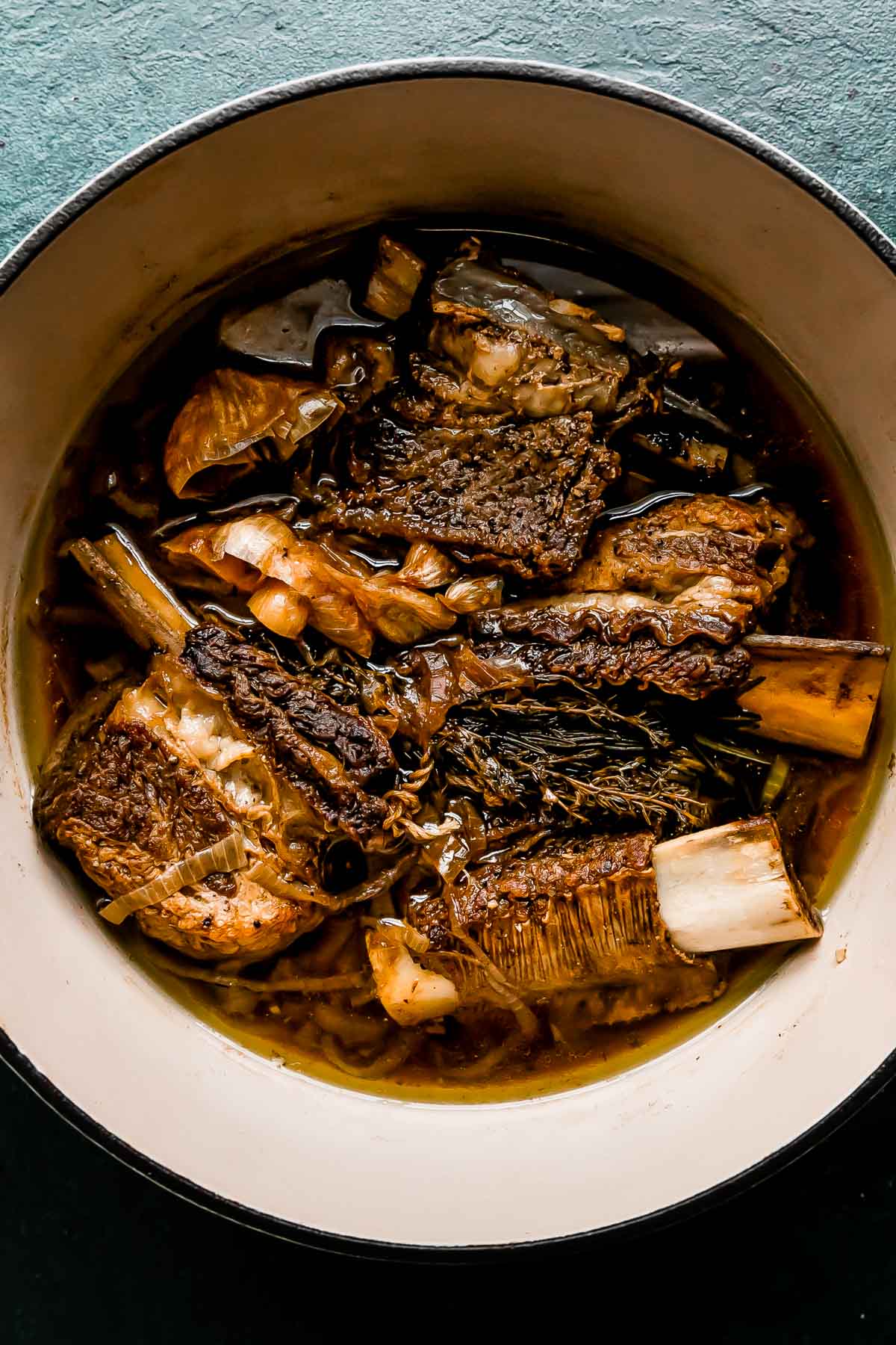 Braised beef short ribs rest inside of a white ceramic pot filled with braising liquid. The pot sits atop a dark green textured surface.