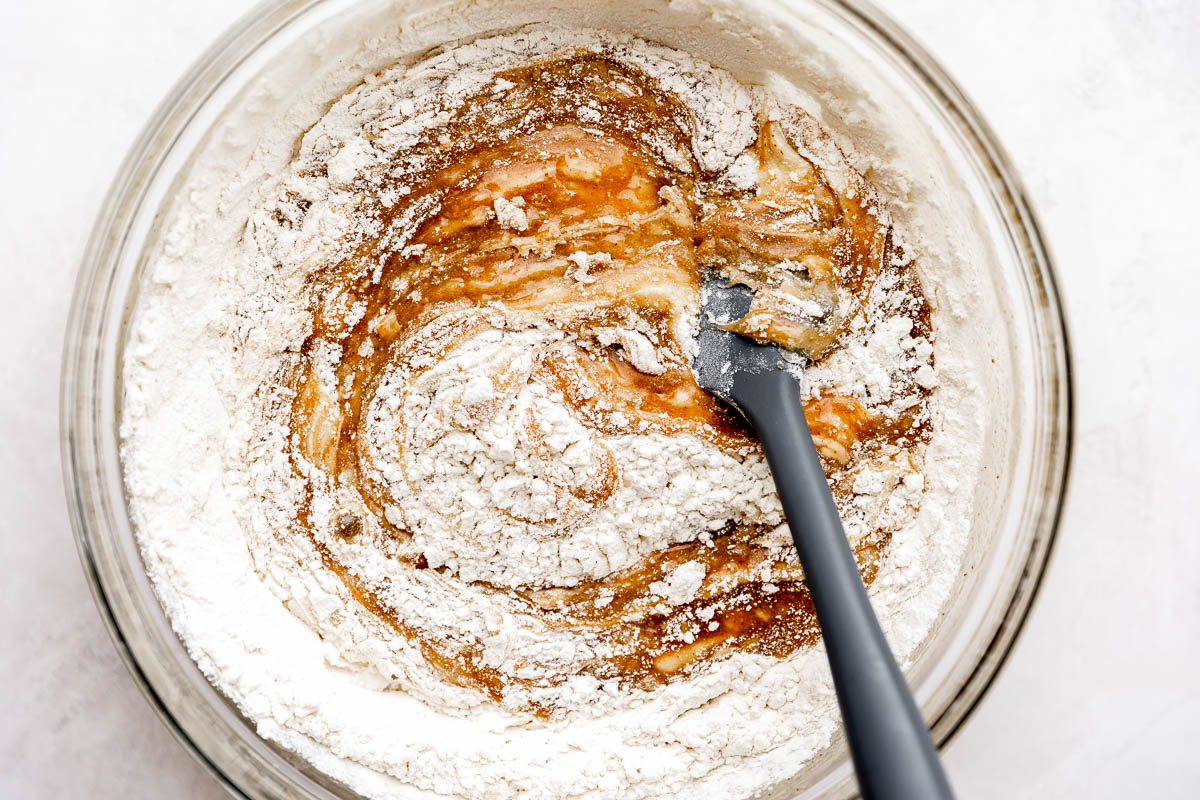 Wet and dry ingredients for brown butter blondies combine inside of a large glass mixing bowl that sits atop a creamy white textured surface. A gray silicone spatula rests inside the bowl.