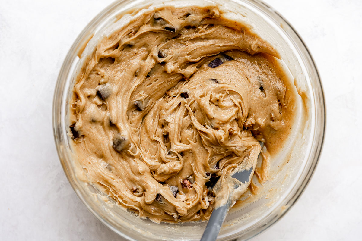 Browned butter blondie batter mixed with pieces of chocolate chunks and chopped walnuts fills a large clear glass mixing bowl. A gray spatula rests inside of the bowl for mixing.