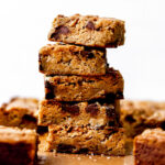 A stack of five brown butter blondies rest atop a piece of parchment paper surrounded by additional browned butter blondies.