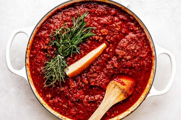 Crushed tomatoes, broth or water, parmesan rind, fresh herbs, and bay leaves are added to a bolognese sauce for Christmas lasagna inside of a large white skillet. The skillet sits atop a creamy white textured surface. A wooden spoon rests inside of the skillet for stirring.
