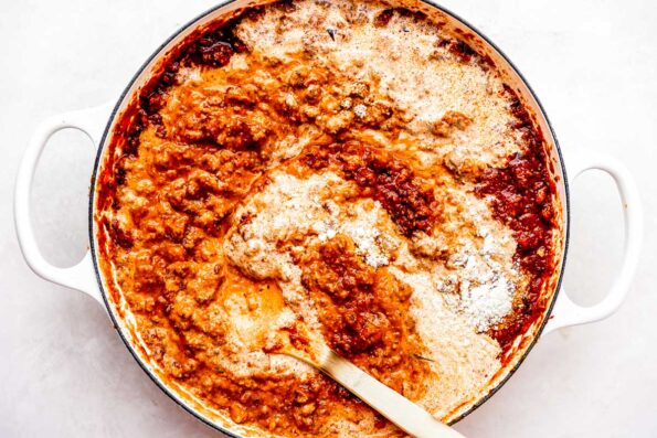 Heavy cream and grated parmesan are added to a homemade bolognese sauce for Christmas lasagna inside of a large white skillet. The skillet sits atop a creamy white textured surface with a wooden spoon resting inside of the skillet for stirring.