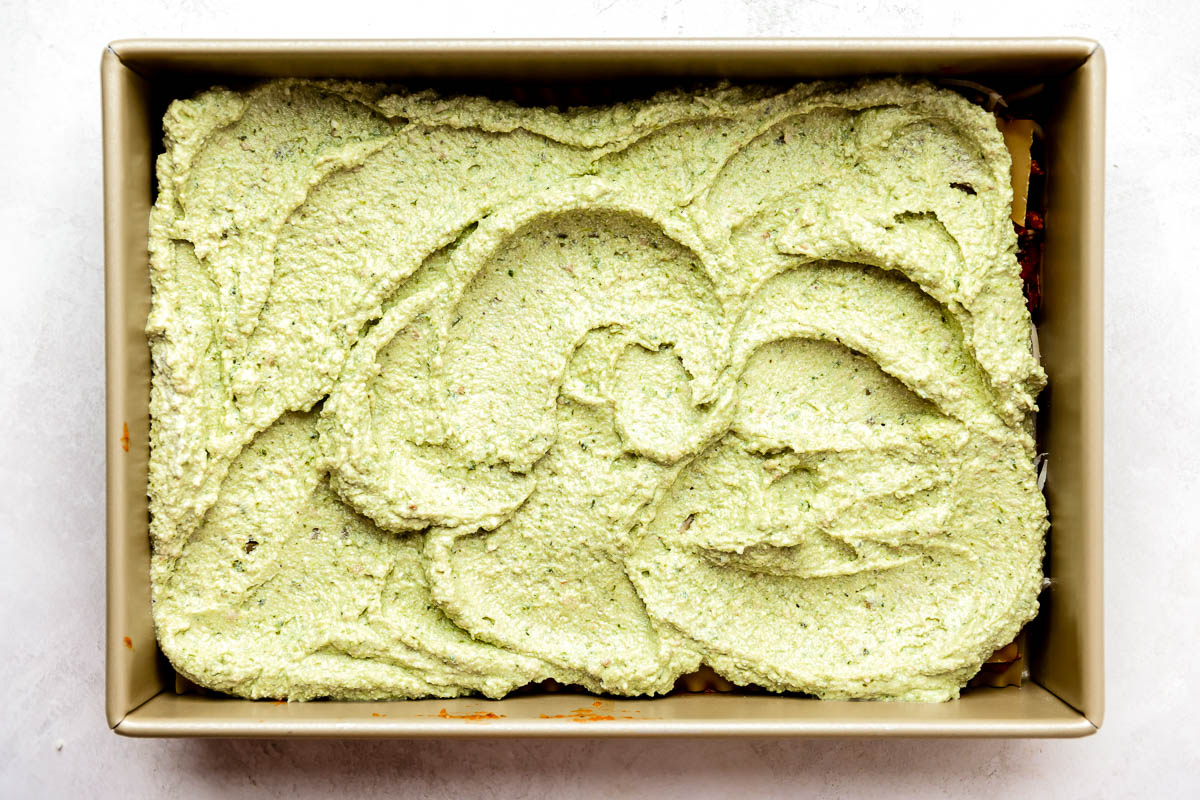 A layer of basil pesto ricotta is spread over top of bolognese sauce and lasagna noodles in a gold 9x13 baking dish to create the second layer of a partially assembled Christmas lasagna. The baking pan sits atop a creamy white textured surface.