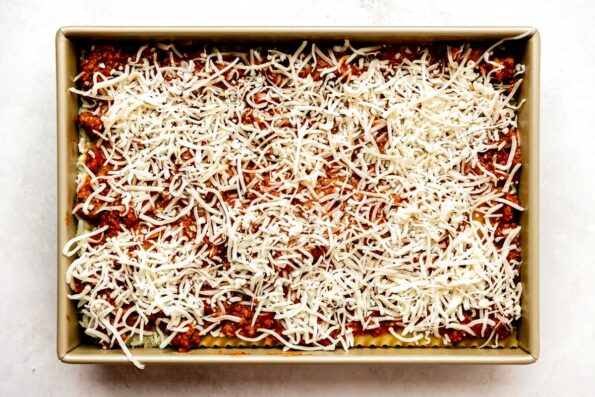 A completely assembled Christmas lasagna fills a gold 9x13 baking dish that sits atop a creamy white textured surface. The top layer of bolognese sprinkled with mozzarella & Italian cheese can be seen on top.