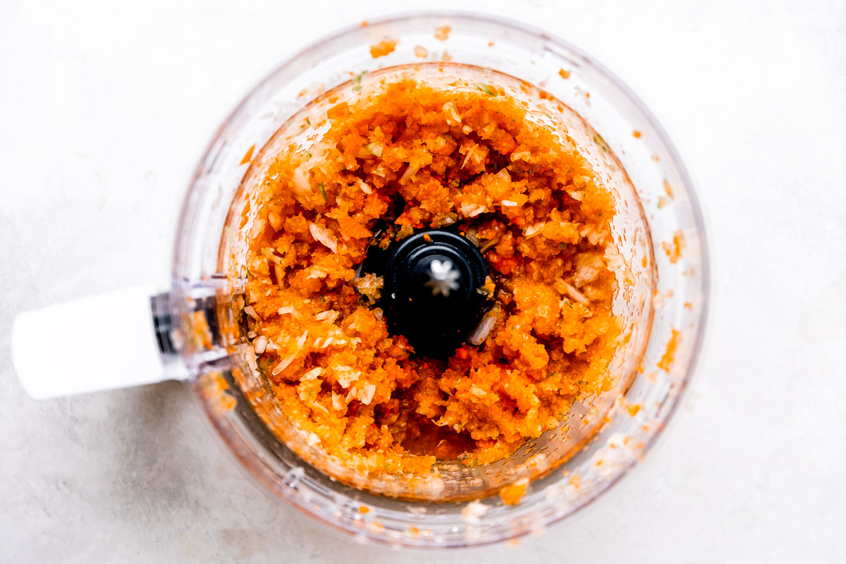 Chopped soffritto veggies fill the bowl of a food processor. The bowl sits atop a creamy white textured surface.