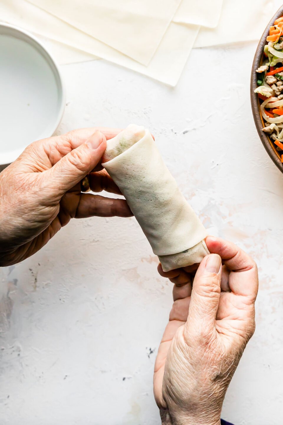 A woman's hands hold a finished assembled pork egg roll over a creamy white textured surface. A yellow ceramic bowl filled with pork egg roll filling, a stack of egg roll wrappers and a small white bowl filled with water rest atop the surface below.