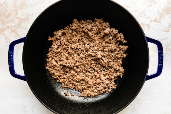 How to make pork egg rolls, step 1: Prepare the pork egg roll filling. Browned ground pork for a pork egg roll recipe fills a large dark blue Staub wok that sits atop a creamy white textured surface.