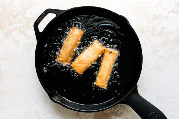Three pork egg rolls shallow fry inside of a large black skillet that sits atop a creamy white textured surface.