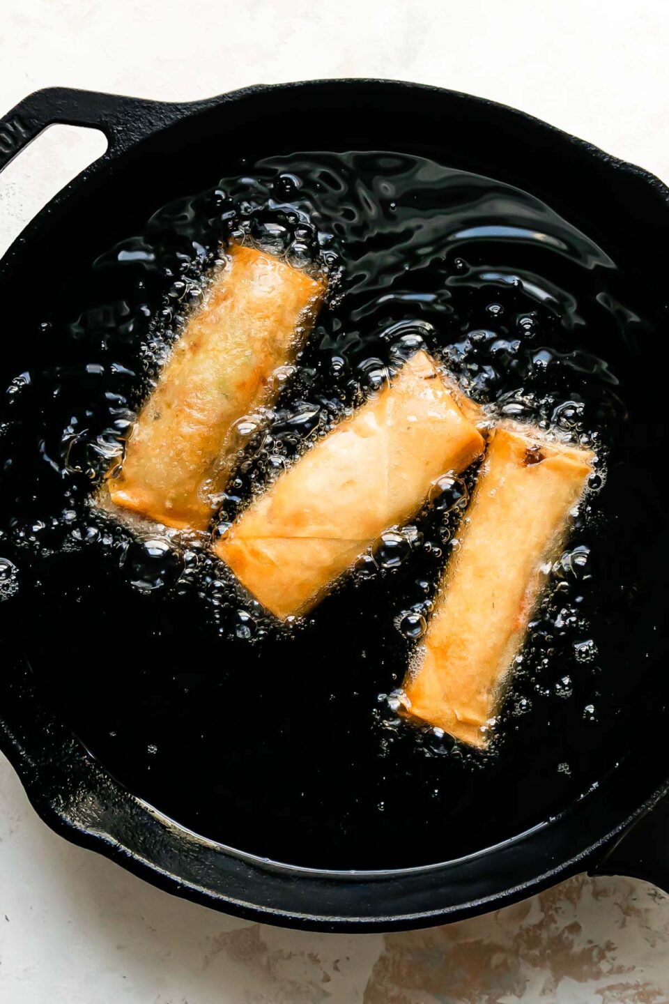 Three pork egg rolls shallow fry inside of a large black skillet that sits atop a creamy white textured surface.