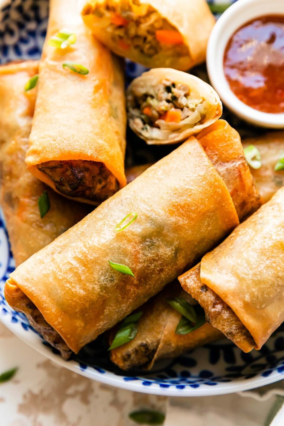 A close up and macro shot of pork egg rolls served inside of a blue and white floral patterned bowl that sits atop a creamy white textured surface. A small white bowl filled with duck sauce rests inside of the bowl. The homemade egg rolls are garnished with sliced green onions and a white linen napkin rests alongside the bowl.