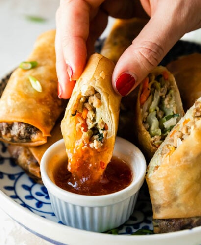A woman's hand holds half of a pork egg roll as it gets dipped inside of a small bowl of duck sauce that sits inside of a blue and white floral patterned bowl filled with more finished egg rolls. The bowl sits atop a creamy white textured surface.