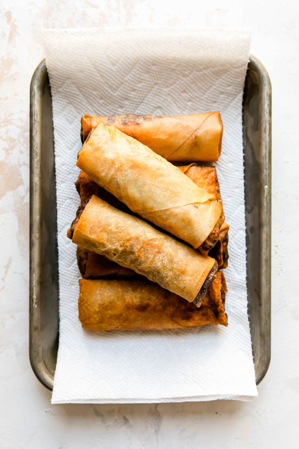 Fried pork egg rolls sit atop a small metal baking sheet pan lined with parchment paper. The sheet pan sits atop a creamy white textured surface.