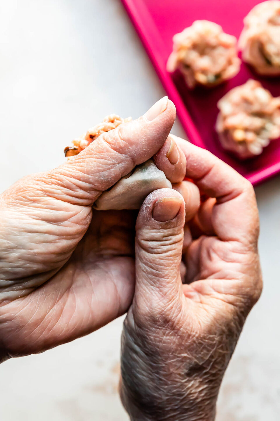 A woman's hands form a pork shumai dumpling over a creamy white textured surface. A pink baking sheet filled with a few assembled shumai dumplings rests atop the surface.