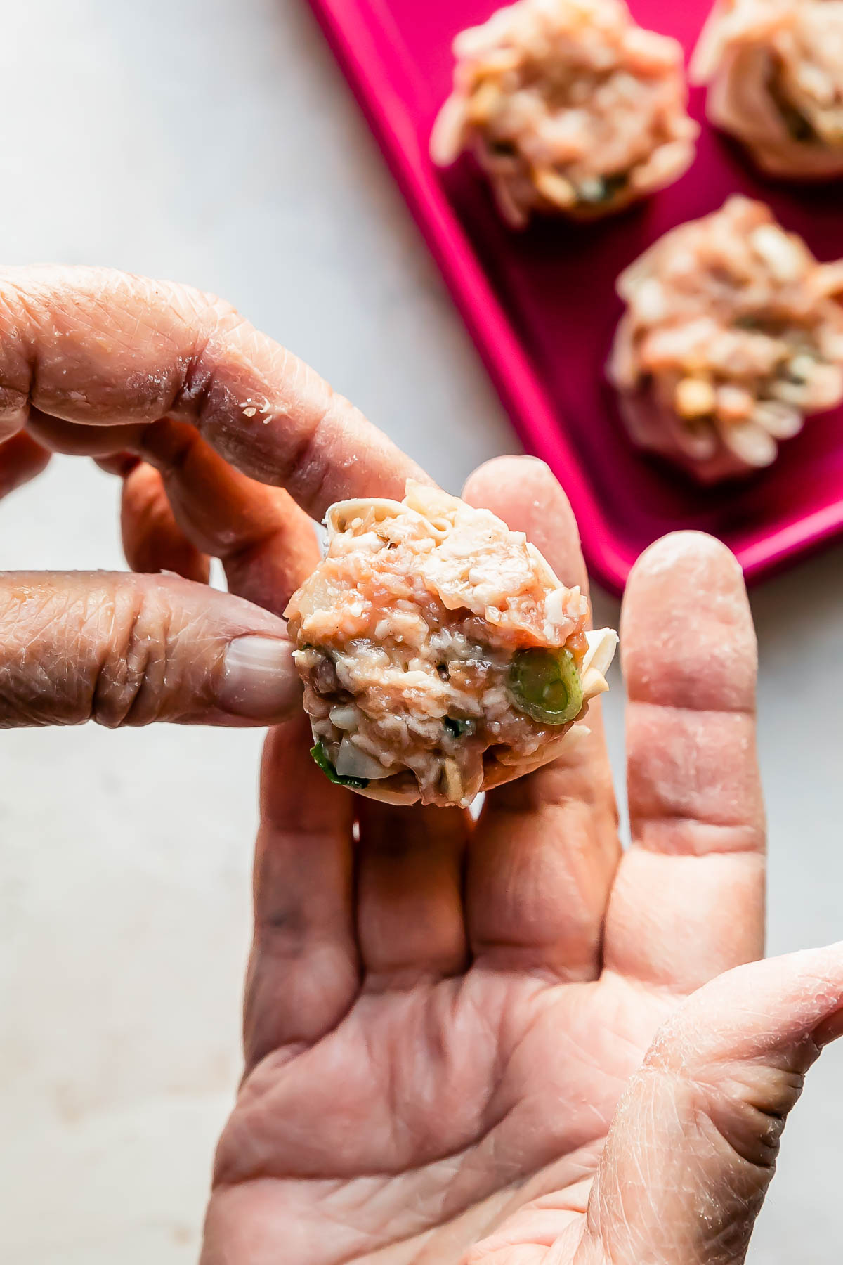 A woman's hands form a shumai dumpling into a purse shape over a creamy white textured surface. A pink baking sheet filled with a few assembled shumai dumplings rests atop the surface.