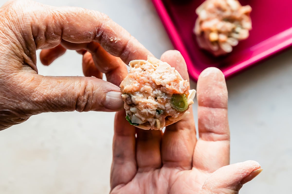 A woman's hands hold an assembled shuami dumpling over a creamy white textured surface. A pink baking sheet filled with a few assembled shumai dumplings rests atop the surface.
