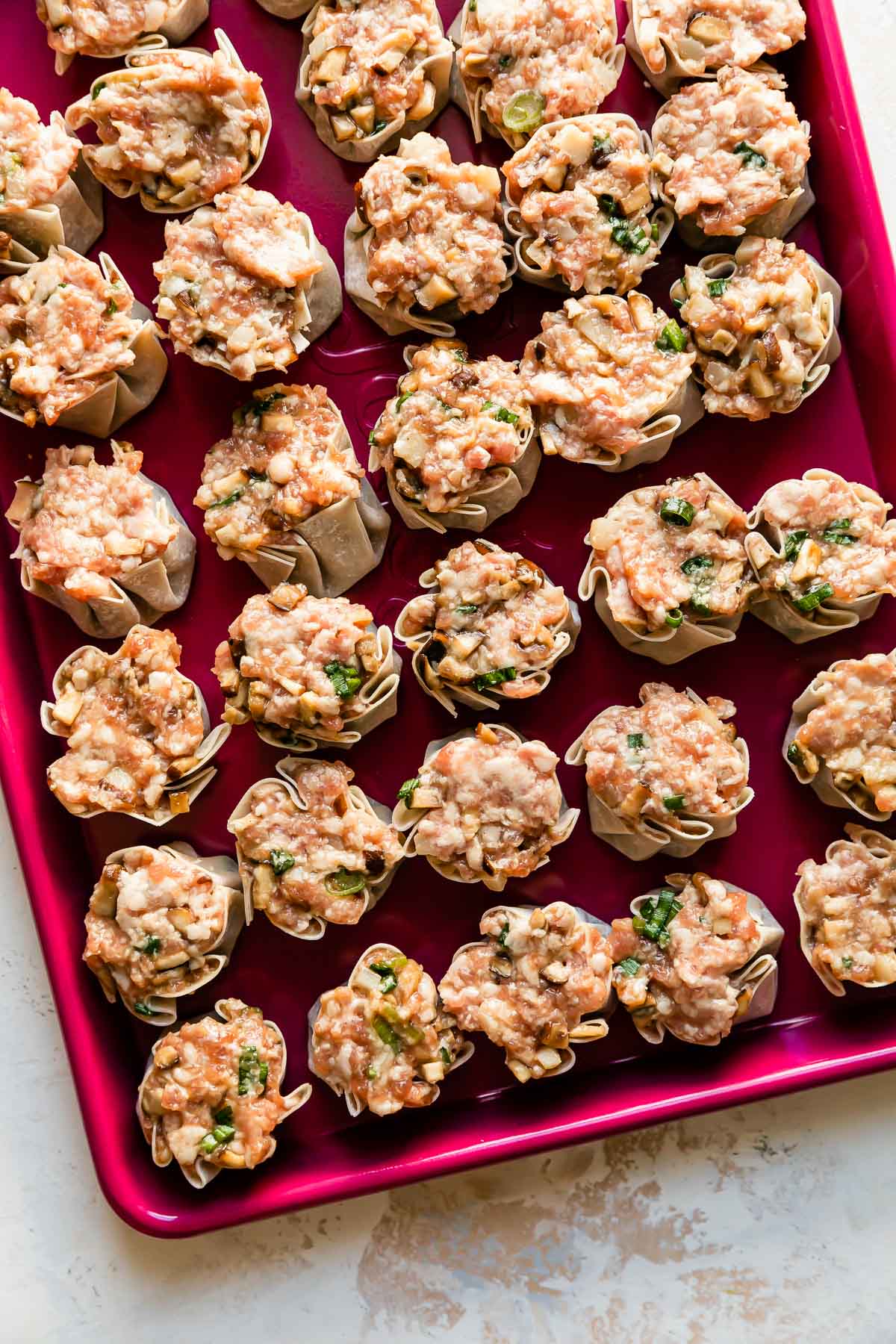 A pink baking sheet is filled with pork shumai dumplings. The baking sheet pan rests atop a creamy white textured surface.