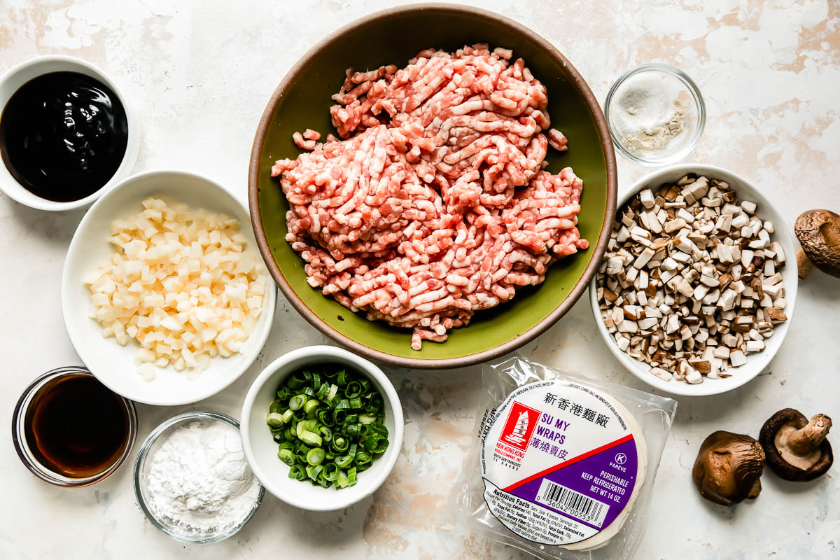 Pork shumai ingredients are arranged atop a creamy white textured surface: ground pork, cornstarch slurry, sesame oil, green onions, dried shiitake mushrooms, water chestnuts, oyster sauce, soy sauce, granulated sugar, white pepper, and round wonton wrappers.