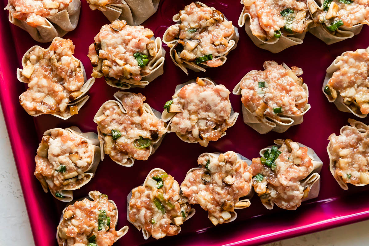 A pink baking sheet is filled with pork shumai dumplings. The baking sheet pan rests atop a creamy white textured surface.