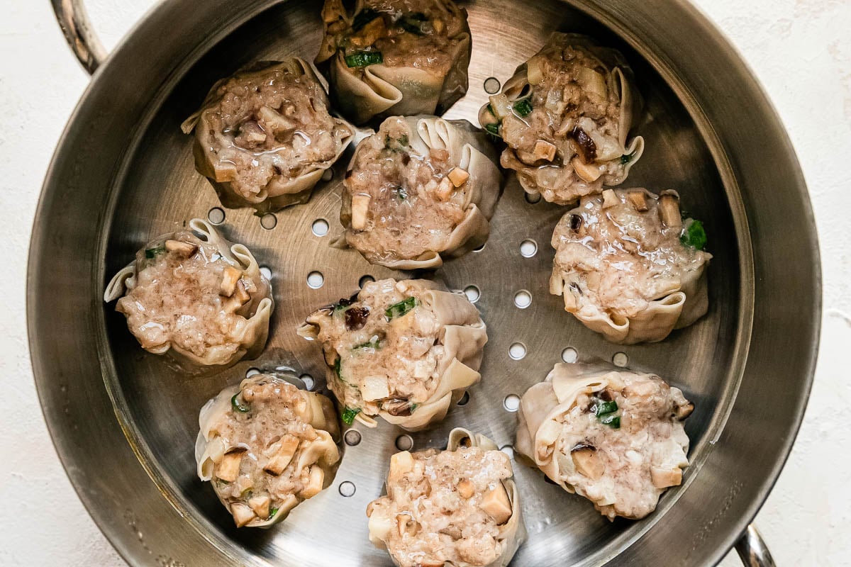 Ten steamed shumai dumplings rest inside of a metal steamer basket that sits atop a creamy white textured surface.