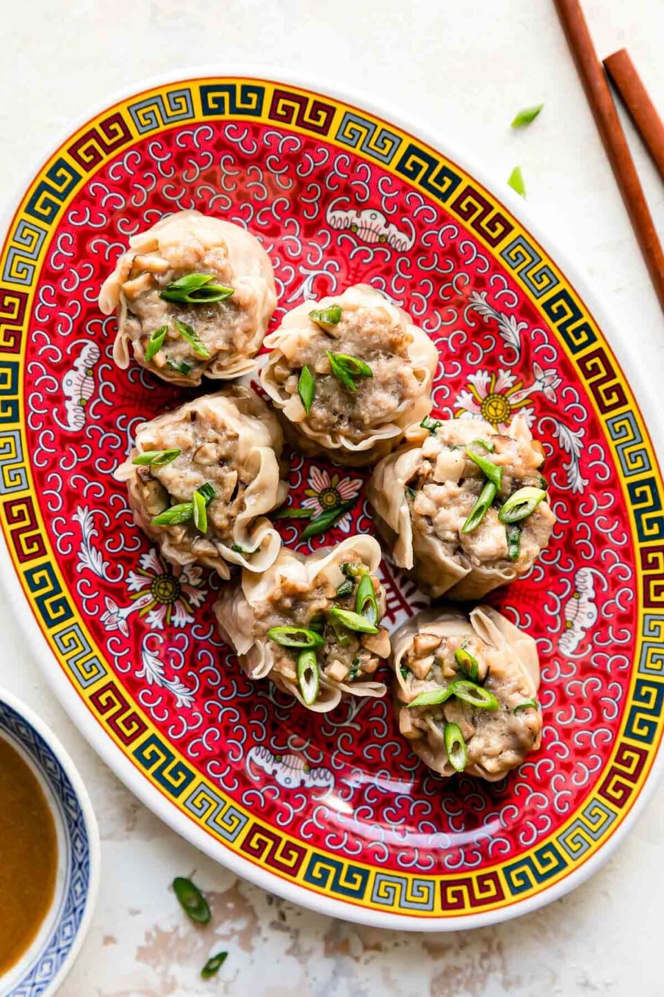 Six pork shumai dumplings rest atop a colorful platter that sits atop a creamy white textured surface. The pork shumai dumplings have been garnished with sliced green onions. A pair of wooden chopsticks rest alongside the platter and a small blue and white bowl filled with siu mai sauce rests alongside the platter.