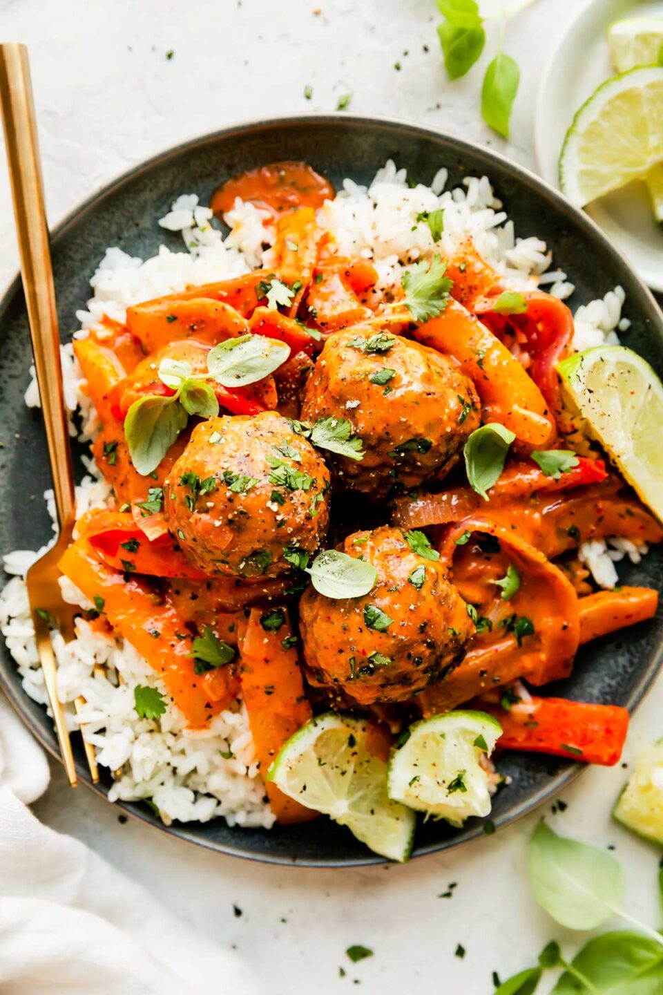 Three red curry meatballs are served atop a plateful of white rice. The blue ceramic plate sits atop a creamy white textured surface with a gold fork resting atop. The plate is surrounded by a small white plate topped with slices of lime, fresh basil leaves, and a white linen napkin.