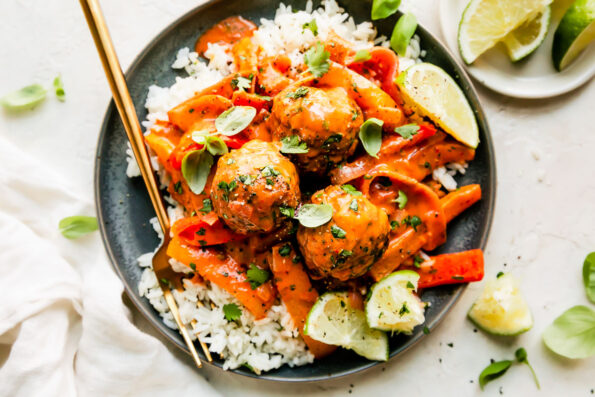 Three red curry meatballs are served atop a plateful of white rice. The blue ceramic plate sits atop a creamy white textured surface with a gold fork resting atop. The plate is surrounded by a small white plate topped with slices of lime, fresh basil leaves, and a white linen napkin.