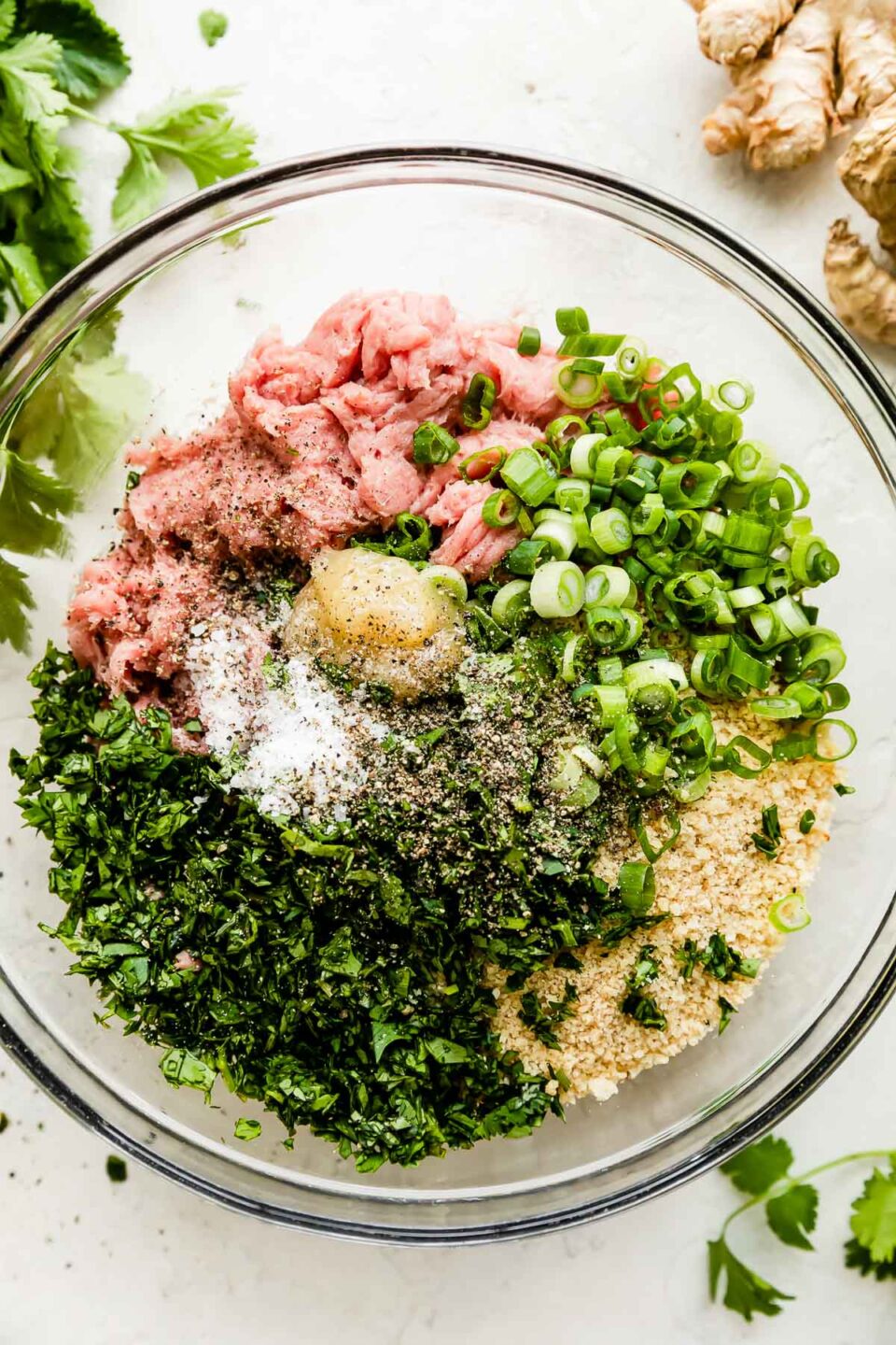 Ground turkey or chicken, green onions, garlic, panko breadcrumbs, cilantro, and ginger are added to large glass mixing bowl that sits atop a creamy white textured surface. The bowl is surrounded by whole fresh ginger and fresh cilantro stems.