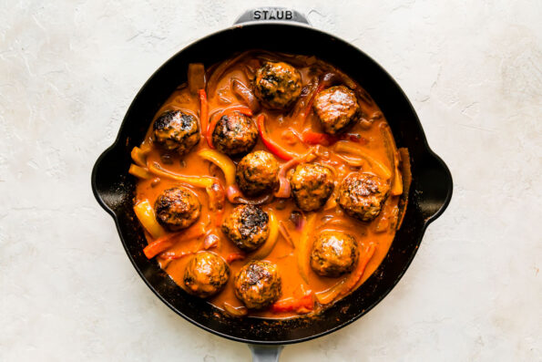 Twelve red curry meatballs simmer in onions, bell peppers and red curry sauce inside of a large gray Staub skillet. The skillet sits atop a creamy white textured surface.