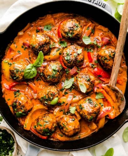 Red curry meatballs fill a large gray Staub cast iron skillet that sits atop a creamy white textured surface. The meatballs have been garnished with fresh basil and cilantro. A wooden serving spoon rests inside of the skillet and the skillet is surrounded by fresh basil leaves, a small wooden pinch bowl filled with chopped cilantro, and a small white plate topped with sliced lime. A light off white linen napkin is tucked underneath the skillet at center.