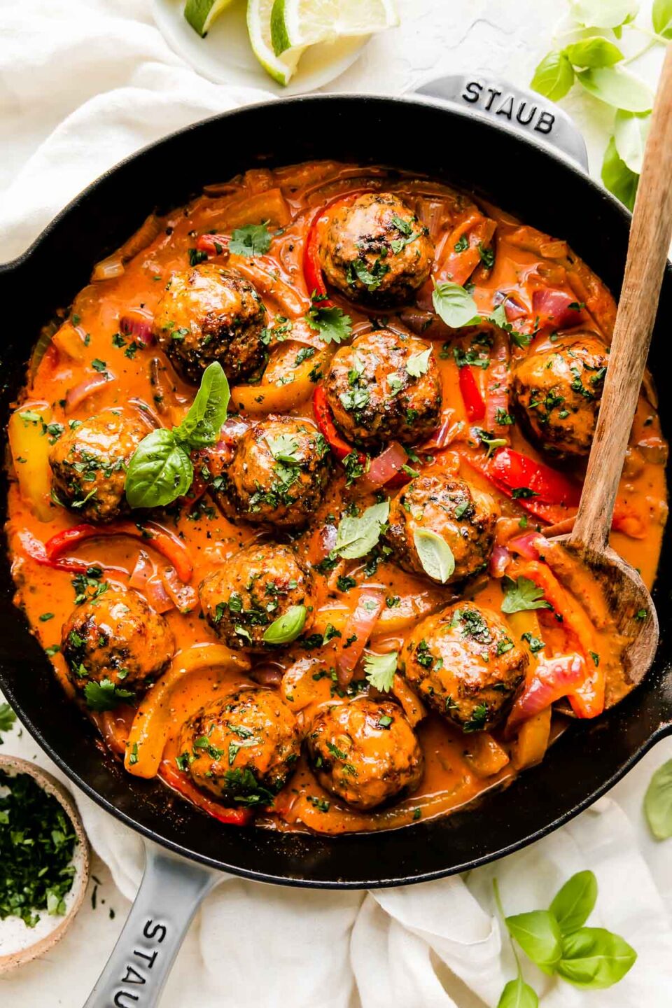 Red curry meatballs fill a large gray Staub cast iron skillet that sits atop a creamy white textured surface. The meatballs have been garnished with fresh basil and cilantro. A wooden serving spoon rests inside of the skillet and the skillet is surrounded by fresh basil leaves, a small wooden pinch bowl filled with chopped cilantro, and a small white plate topped with sliced lime. A light off white linen napkin is tucked underneath the skillet at center.