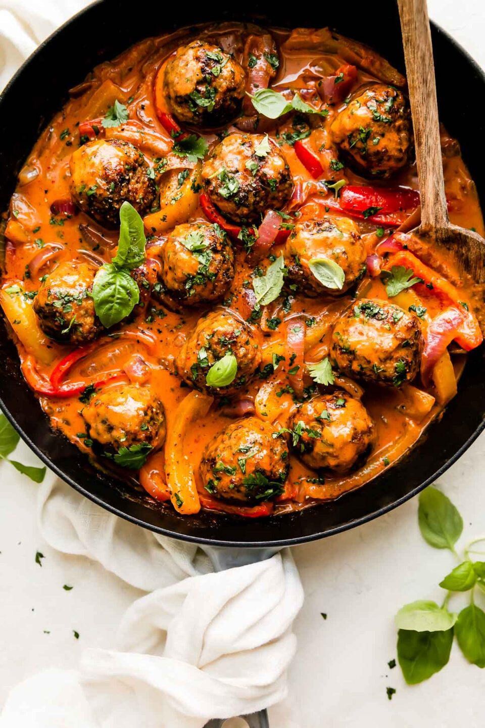 Curry turkey meatballs fill a large gray Staub cast iron skillet that sits atop a creamy white textured surface. The meatballs have been garnished with fresh basil and cilantro. A wooden serving spoon rests inside of the skillet and the skillet is surrounded by fresh basil leaves with a white linen napkin is tucked underneath the skillet and around the skillet's handle.