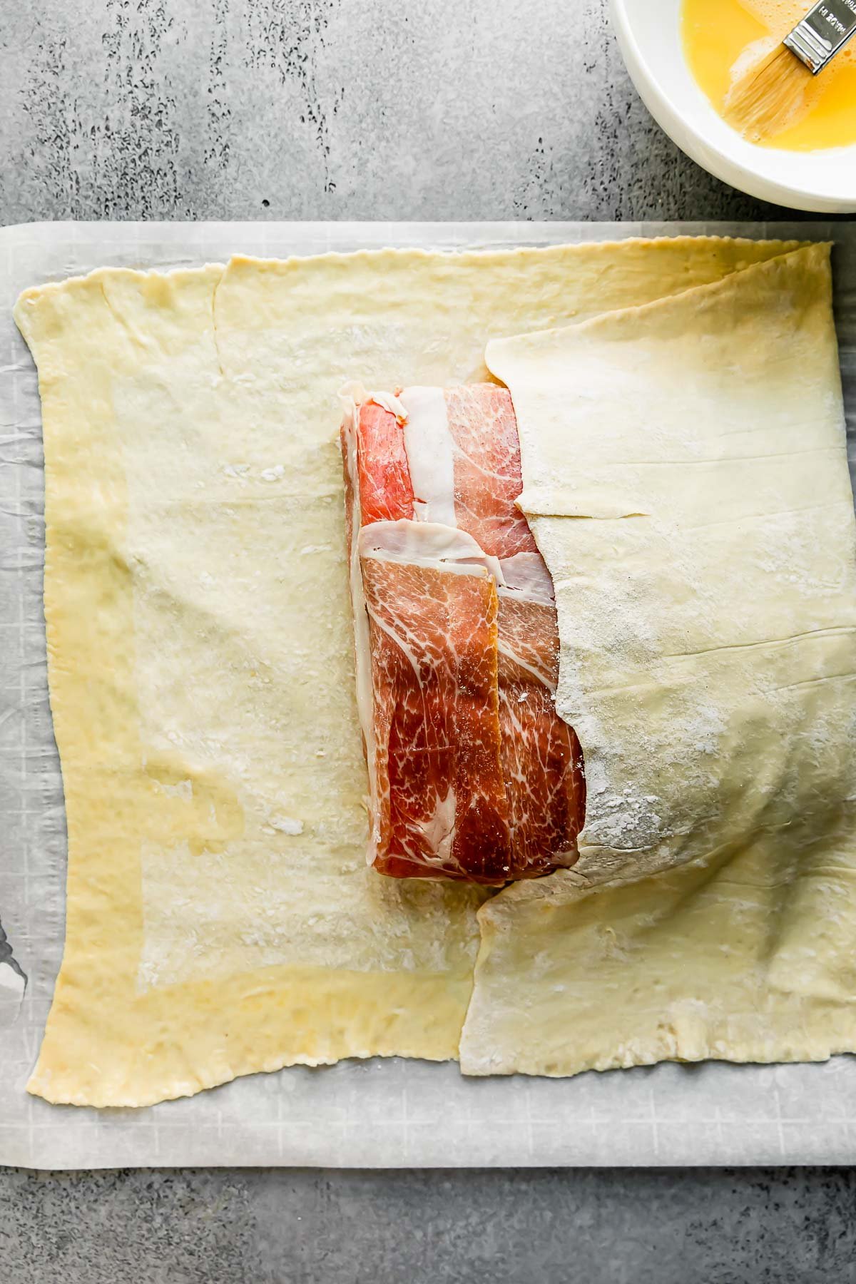 A large rectangular piece of puff pastry is partially folded over and atop of prosciutto-wrapped salmon. The puff pastry & salmon sit atop a large piece of parchment paper that rests atop a light gray textured surface. This is the begining of salmon wellington forming. A small white bowl filled with egg wash with pastry brush resting alongside rests near the salmon en croute.