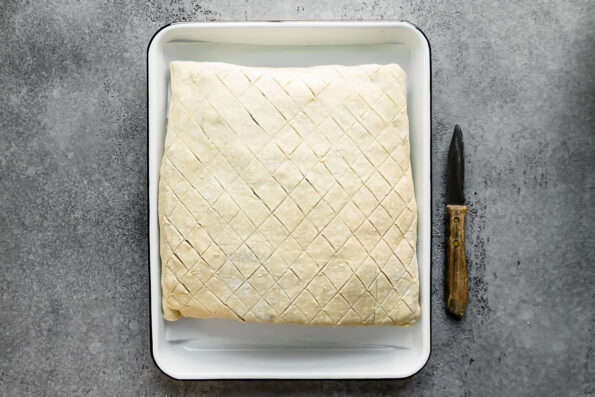 Raw and unbaked salmon wellington rests inside of a white parchment lined baking tray that sits atop a light gray textured surface. The surface of the puff pastry has been scored in a crosshatch pattern and a small paring knife rests alongside the tray.