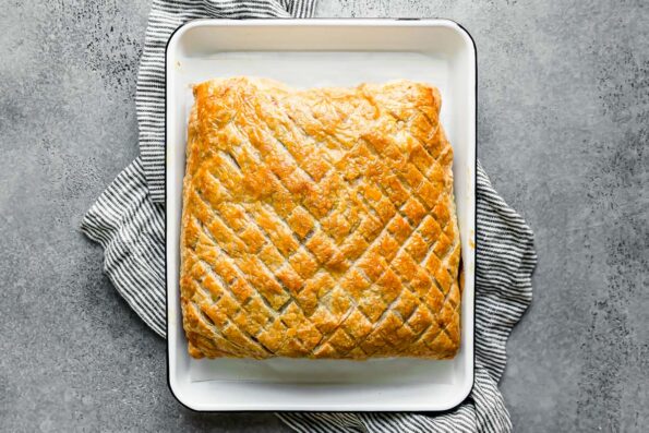 Baked salmon wellington rests inside of a white parchment lined baking tray that sits atop a light gray textured surface. The surface of the puff pastry has been scored in a crosshatch pattern and the puff pastry is golden brown. A blue and white striped linen napkin rests underneath the baking tray at center.