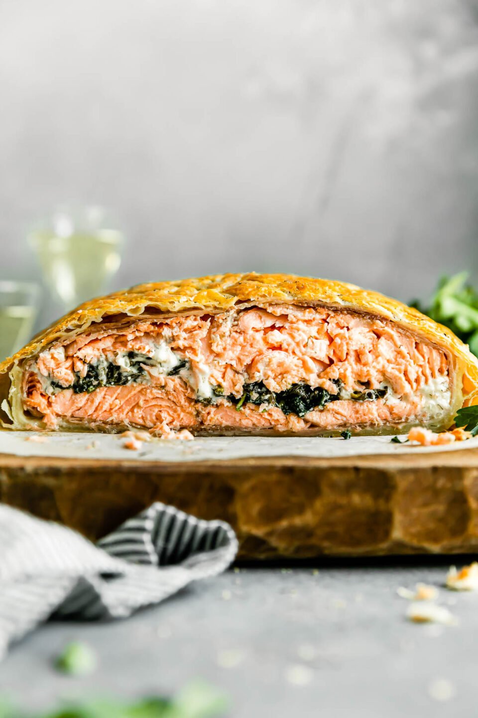 A side angle shot of a baked and sliced cross-section of salmon wellington rests atop a piece of parchment paper that sits atop a natural wood platter. Two glasses of white wine, fresh herbs, and a blue and white striped linen napking surround the salmon en croute at center.