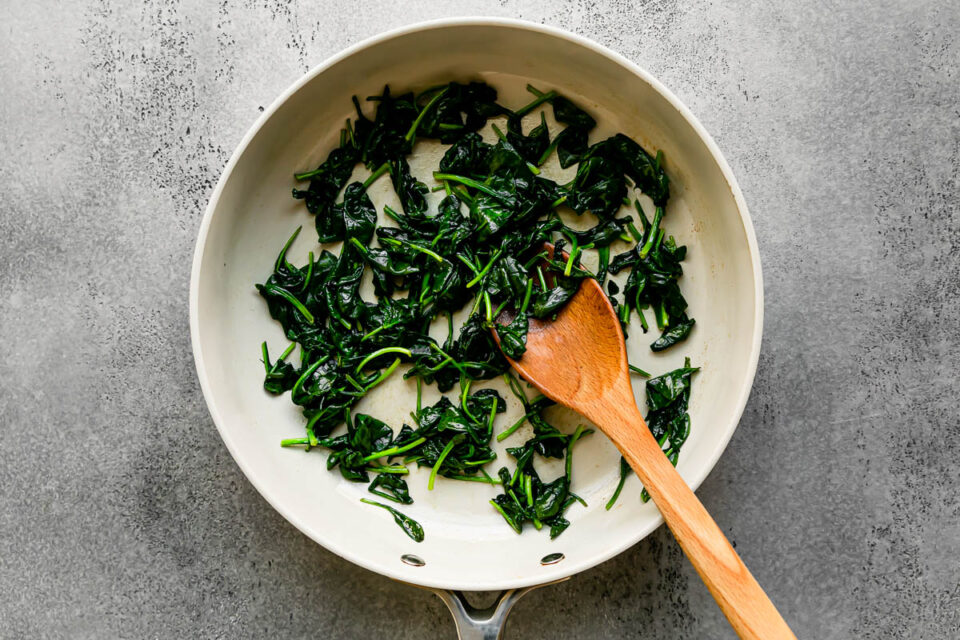 Wilted spinach cooked in melted butter in a light colored skillet sits atop a light gray textured surface. A wooden spoon rests inside of the skillet for stirring.