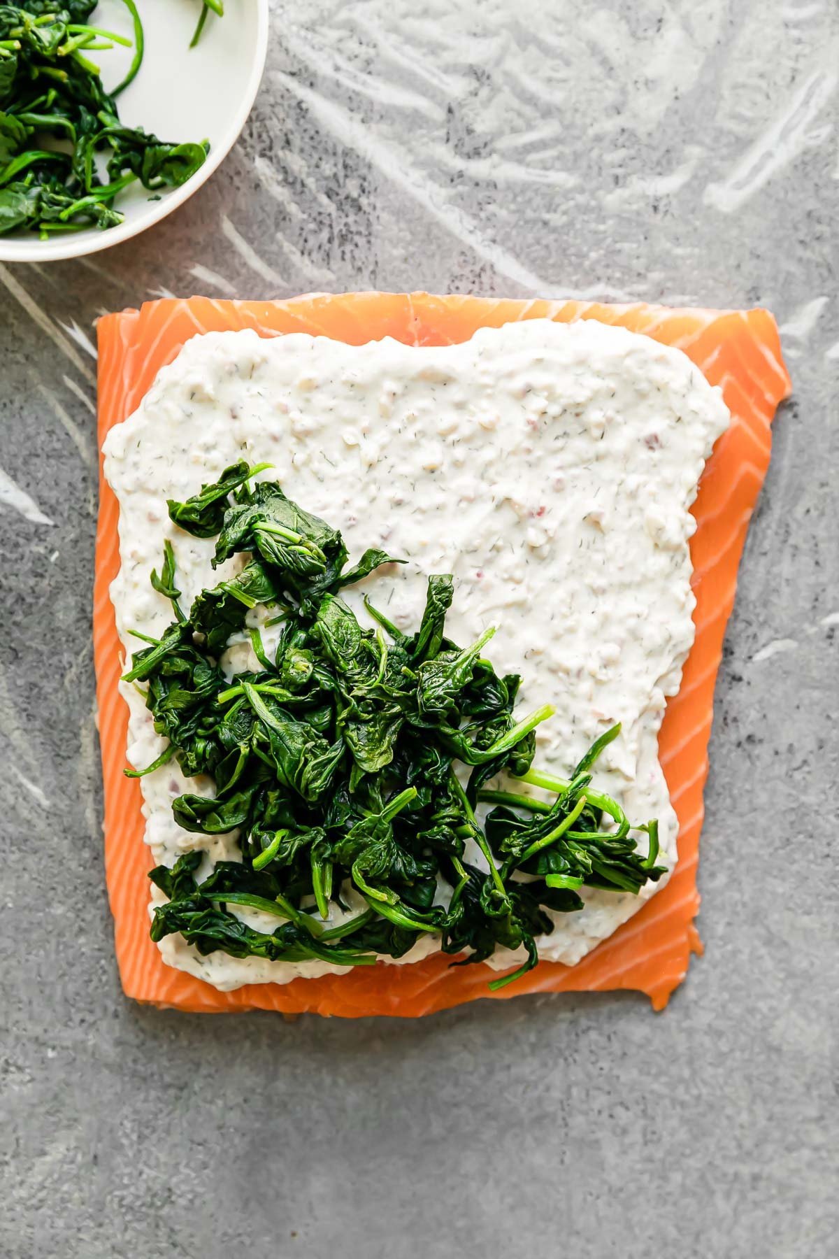 A single salmon fillet sits atop a large piece of plastic wrap. The salmon fillet has been layered with a garlic & dill cream cheese spread and wilted spinach as salmon wellington begins to be built. A small white bowl filled with wilted spinach rests alonside the salmon & both the salmon & the bowl sit atop a light gray textured surface.