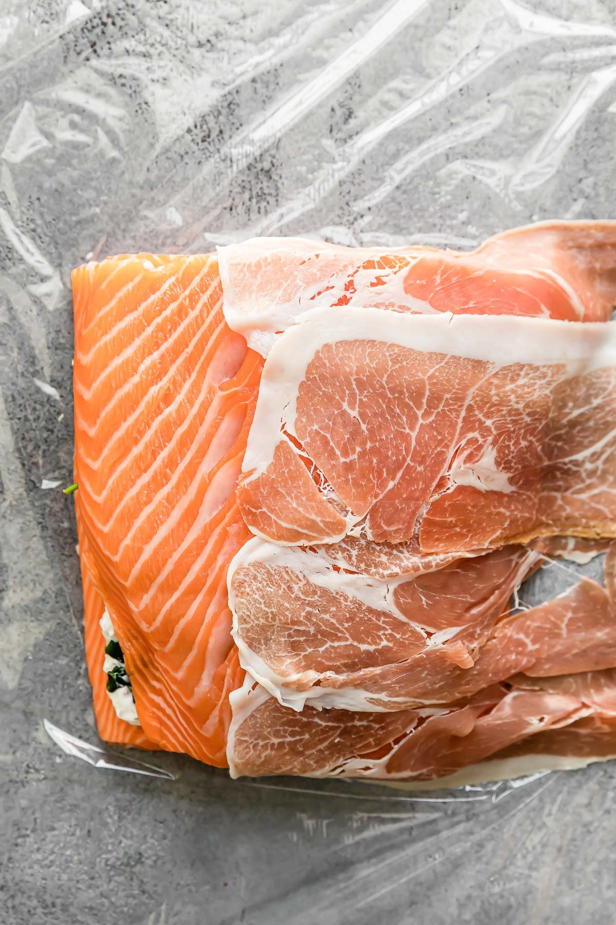 Layered salmon fillets sit atop a large piece of plastic wrap. Pieces of prosciutto are placed over top of the fillets, overlapping eachother to begin covering completely.