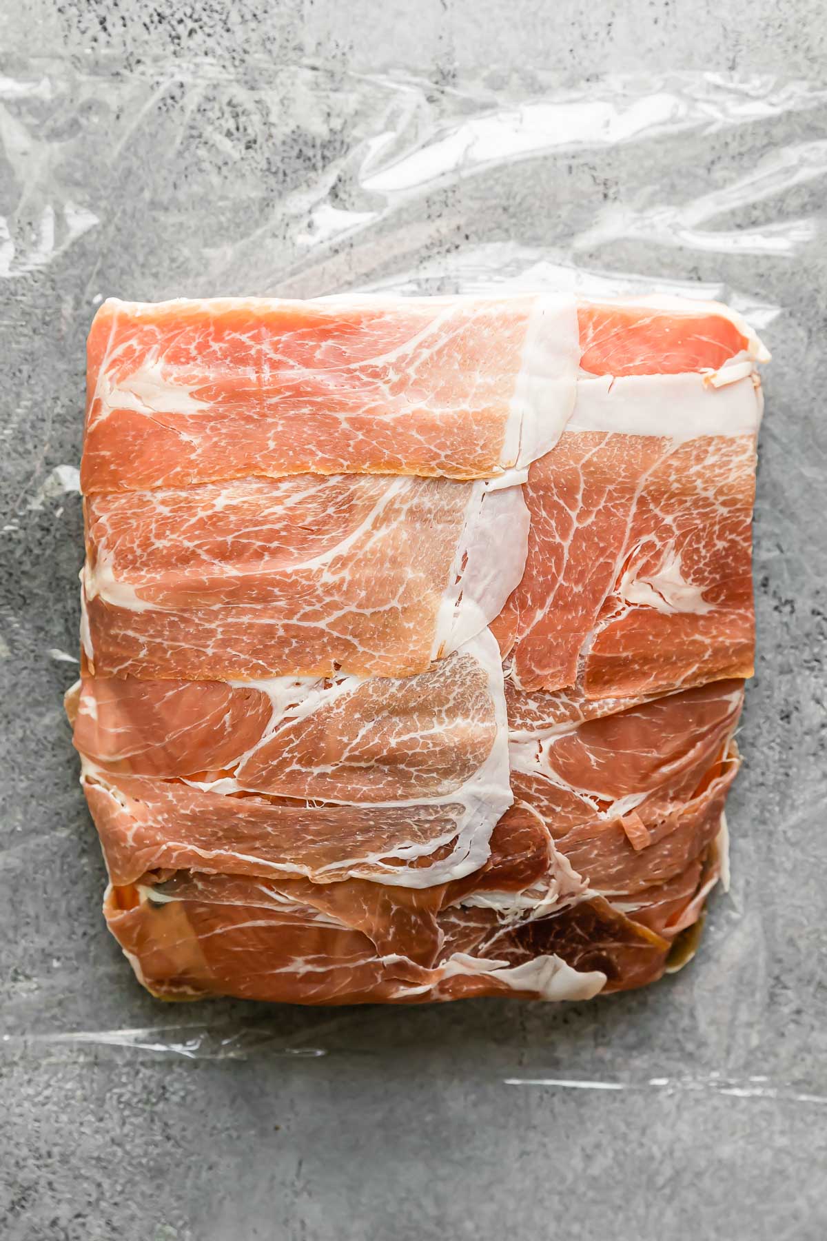 Layered salmon fillets sit atop a large piece of plastic wrap. Pieces of prosciutto are placed over top of the fillets, overlapping eachother to cover completely.