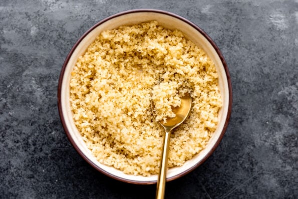 Crispy breadcrumbs are mixed inside of a small ceramic bowl that sits atop a dark gray textured surface. A gold spoon rests inside of the bowl for mixing.