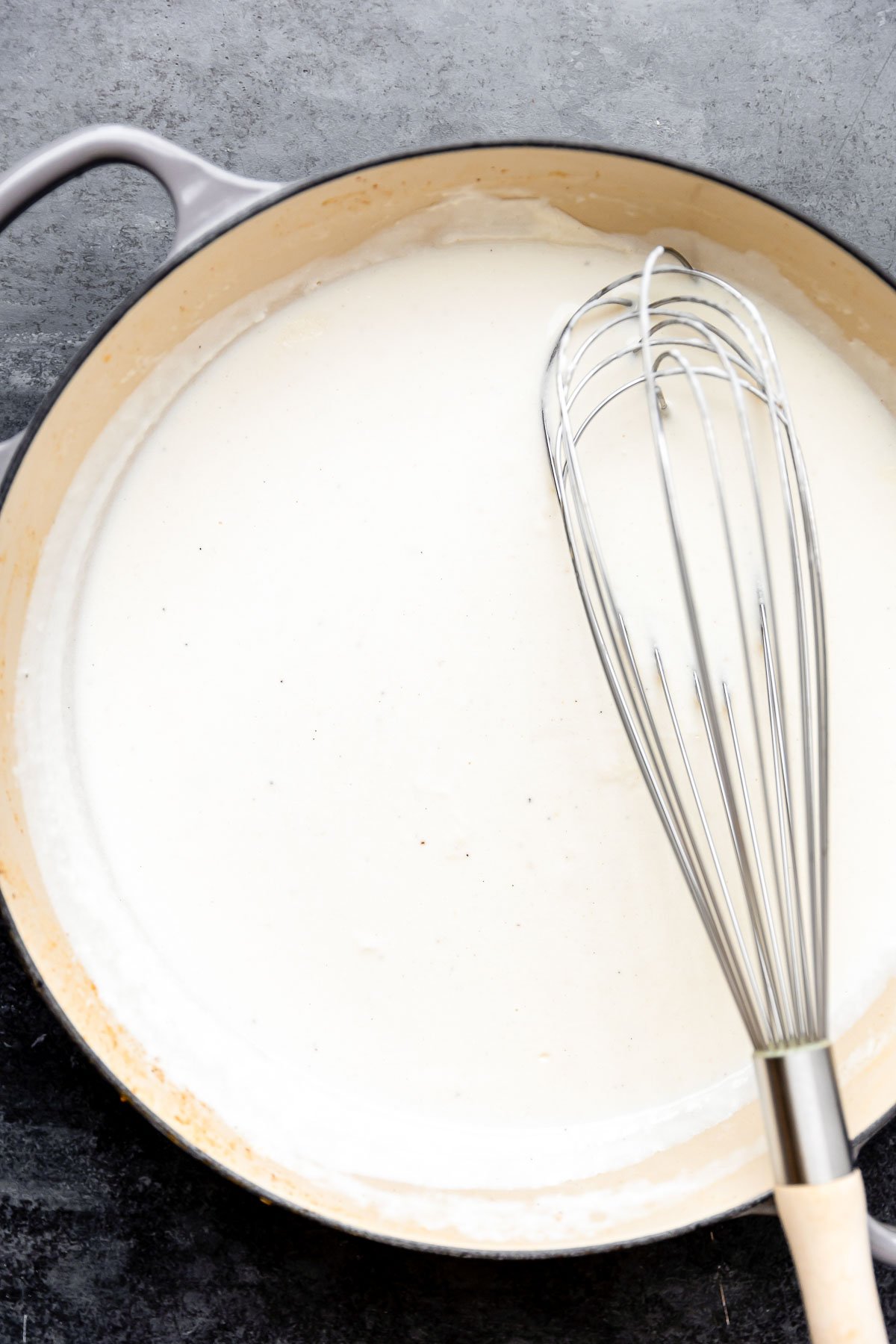 Seafood béchamel sauce for seafood stuffed shells fills a large gray skillet that sits atop a dark gray textured surface. A metal whisk rests atop the skillet for mixing.