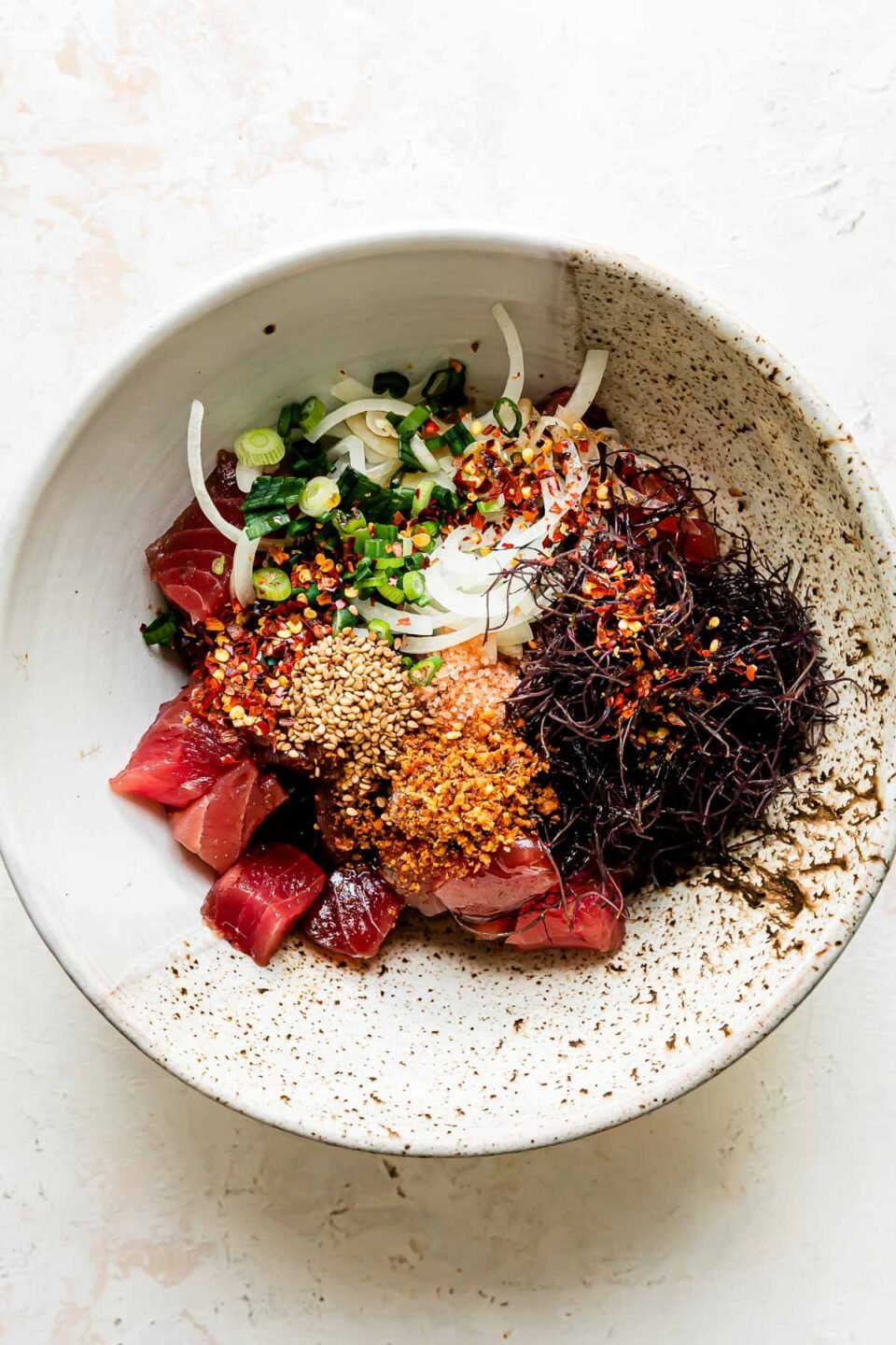 Tuna poke components are arranged in a large ceramic bowl that sits atop a creamy white textured surface: ahi tuna, sweet onion, green onions, ogo or limu, shoyu, toasted sesame oil, Hawaiian sea salt, Inamona, toasted sesame seeds, and crushed red pepper.